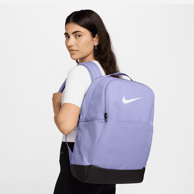 Eine Frau mit langen dunklen Haaren in einem weißen Hemd und einer schwarzen Hose posiert mit dem Schulrucksack Nike, 5 Klasse und Gymnasium, Schulranzen Mädchen Jungen, Oberstufe und Teens auf ihrer Schulter vor einem schlichten weißen Hintergrund.
